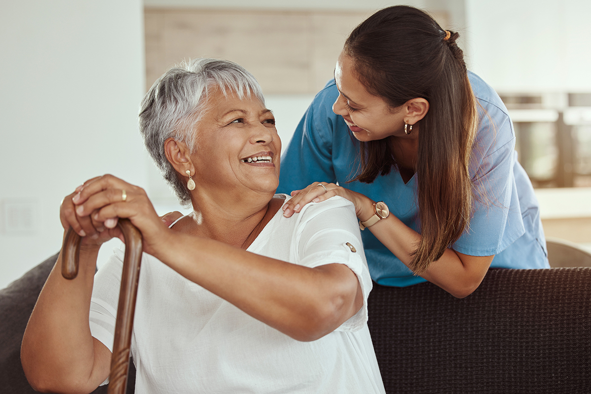 Happy, relax and senior woman with caregiver smile while sitting on a living room sofa in a nursing home. Support, help and professional nurse or healthcare worker helping elderly lady or patient